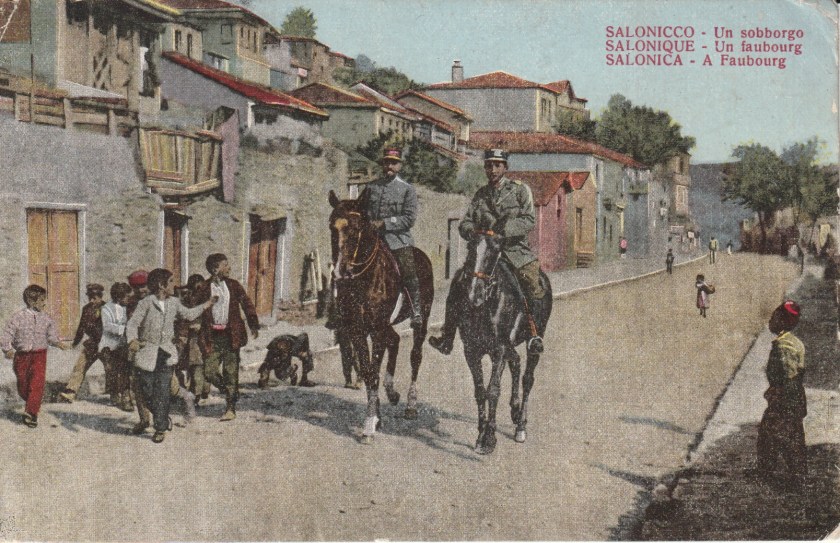 Hand-tinted postcard showing a suburb of Salonika, with children following two mounted soldiers or gendarmes.